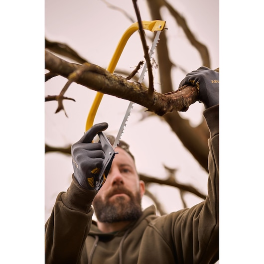 Photograph of person using STANLEY 1-20-447 bow saw to cut tree branch, wearing gloves and brown jacket outdoors.