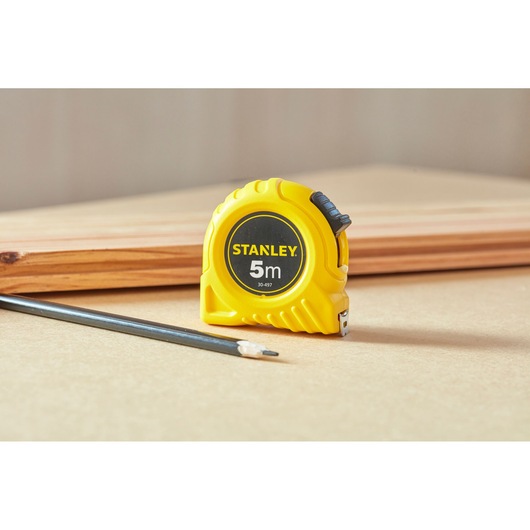 Stanley 1-30-497 tape measure in yellow casing, placed beside pencil and wood, product photography.