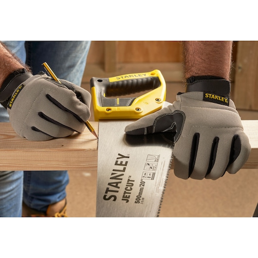 STANLEY 2-15-288 hand saw and yellow stapler on wood, person with gloves marking cut line, workshop background.