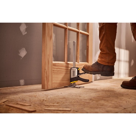 STANLEY FMHT83550-1 door installation clamp in use on a wooden door, worker using foot to operate tool.